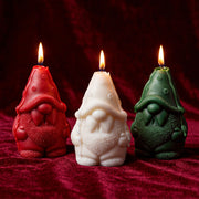 Trio of lit gnome pillar candles in red, white and green on deep red velvet fabric background - Boowan Nicole