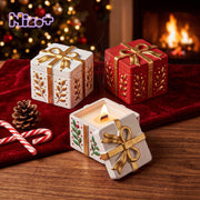 Assorted finished Holiday Gift Box Candle Jar candle vessels in red and white with gold bow‑leaf details on red velvet against Christmas fireplace background - Boowan Nicole