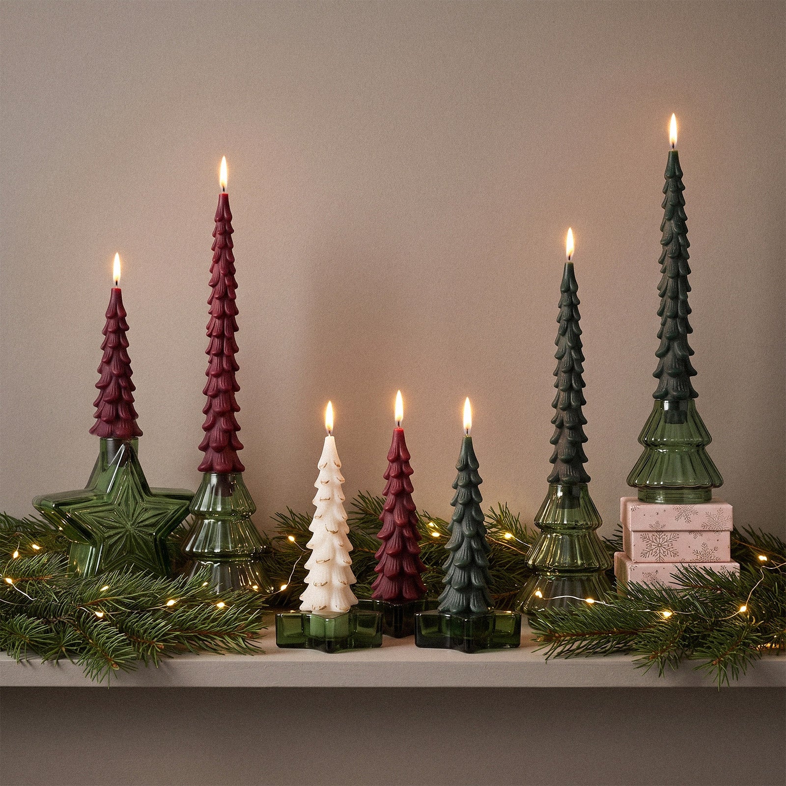 Assorted sizes of winter pine tree taper candles lit on shelf with festive evergreen decor - Boowan Nicole