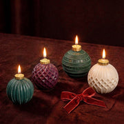 Four textured spherical wax candles with gold top caps and lit flames, arranged on a dark brown velvet surface with a red bow. The candles are in teal, purple, green, and cream colors.