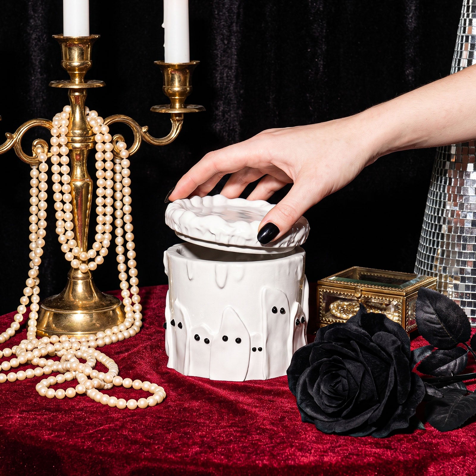 Hand lifting the lid of white ghost cake candle jar beside pearl necklace, golden candlestick and black rose for gothic display - Boowan Nicole