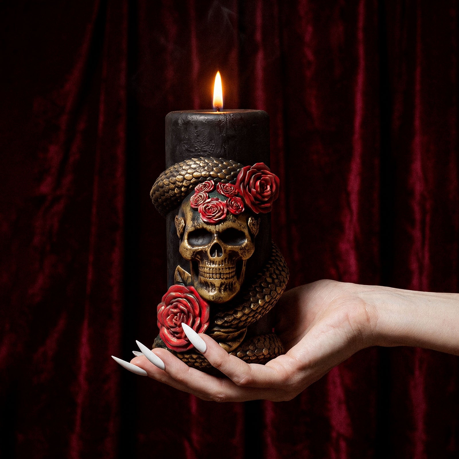 Hand holding tall lit Venom & Roses skull snake rose pillar candle against burgundy velvet curtain background - Boowan Nicole