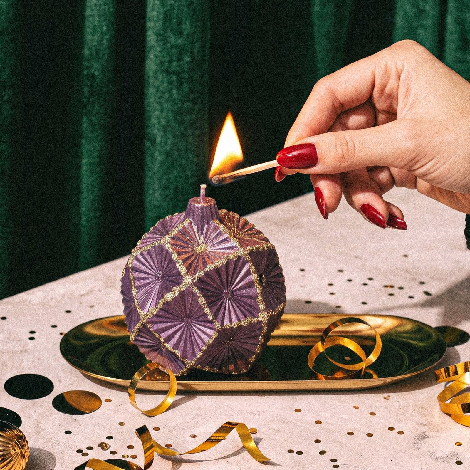 Hand lighting purple geometric bauble candle placed on golden metal tray with festive confetti decor - Boowan Nicole