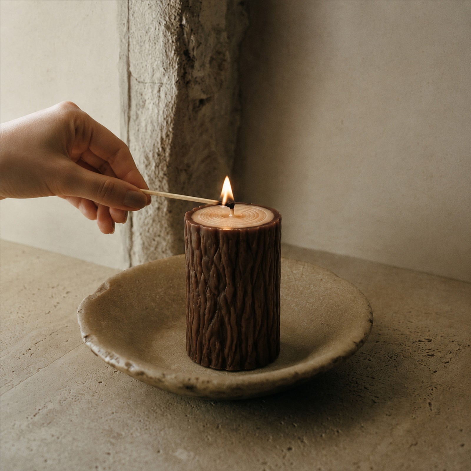 Hand lighting dark tree stump pillar candle with a match, rustic minimalist stone plate styling - Boowan Nicole
