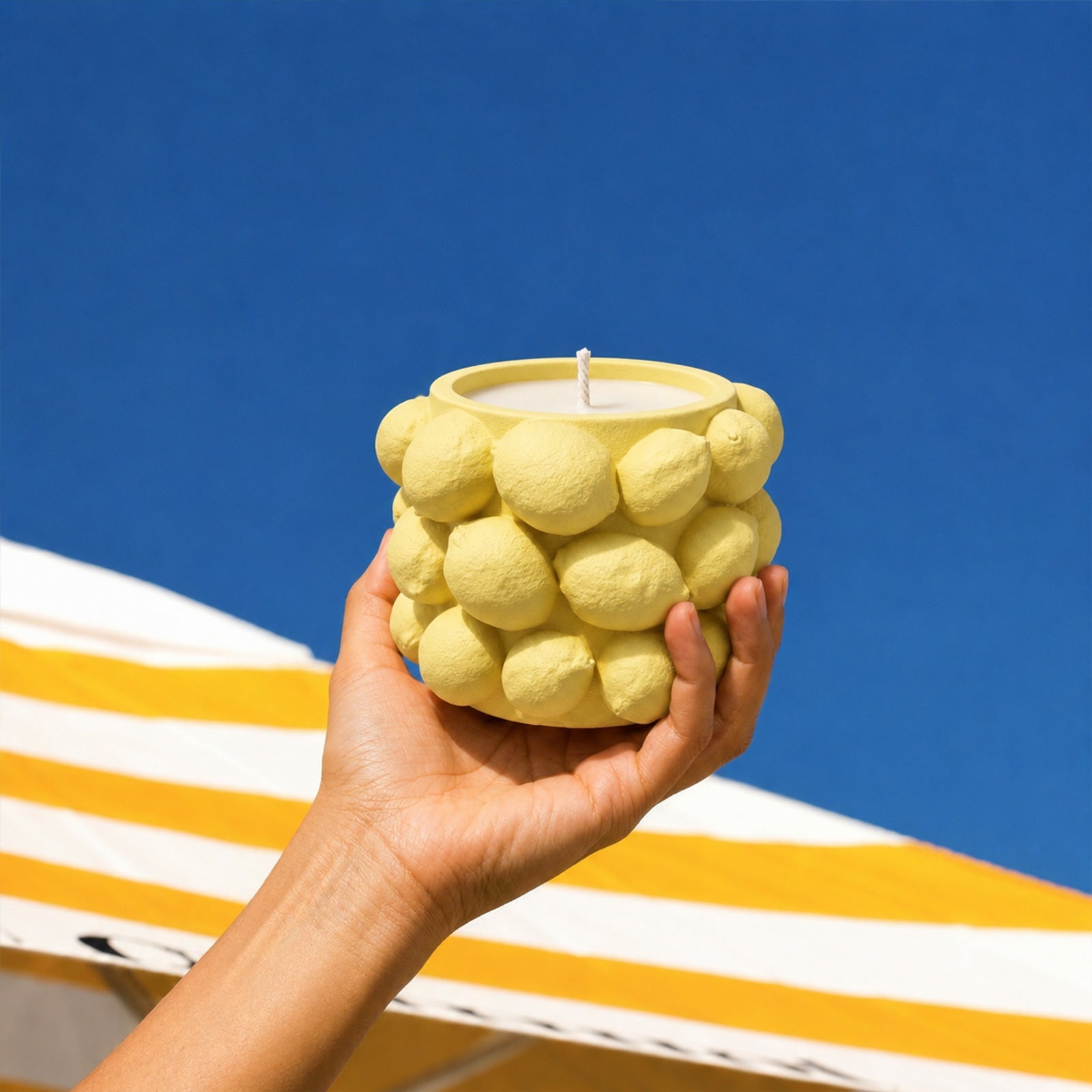 Hand holding yellow Lemon Drops candle jar against blue sky and striped awning - Boowan Nicole