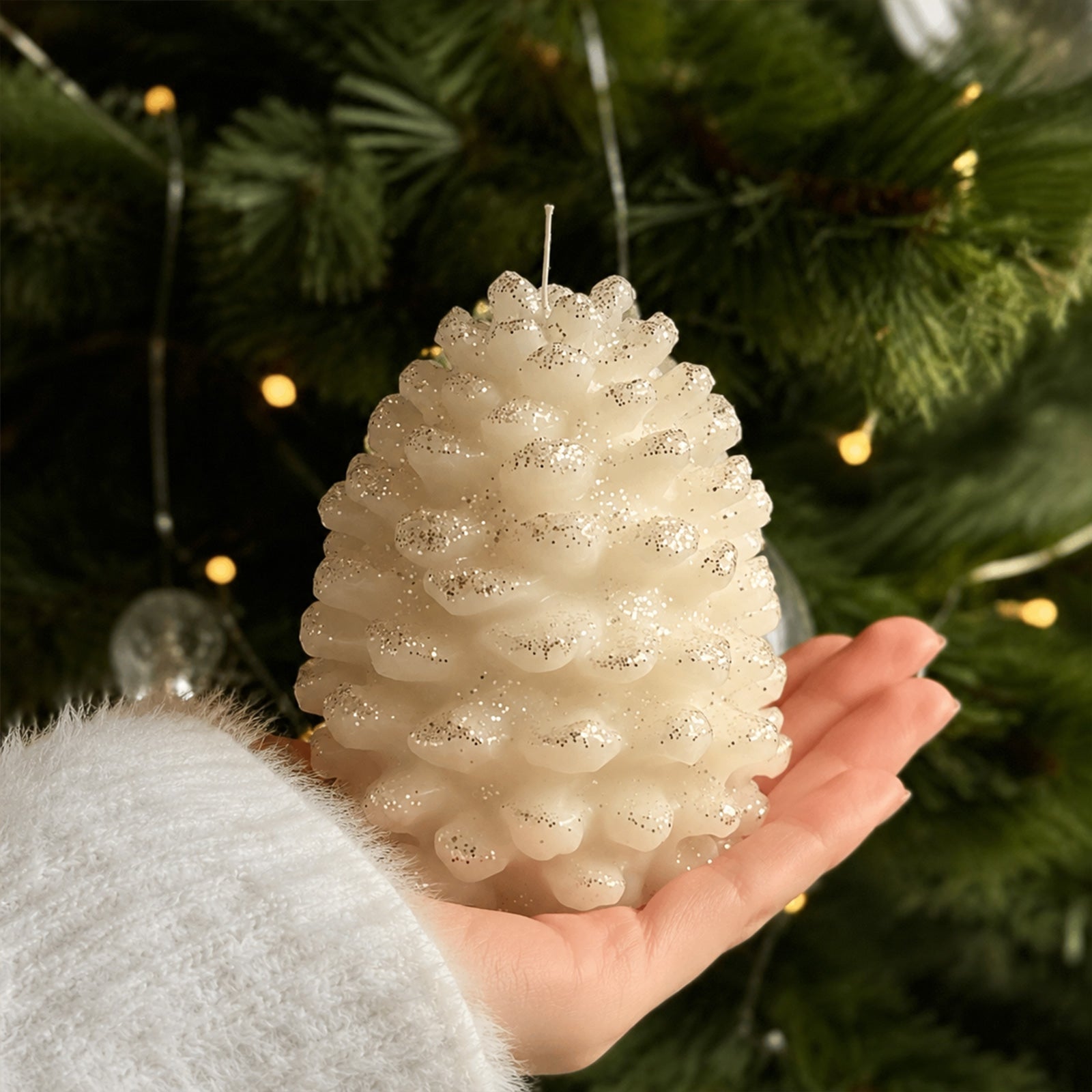 Hand holding cream glitter winter pinecone pillar candle in front of decorated Christmas tree lifestyle shot - Boowan Nicole