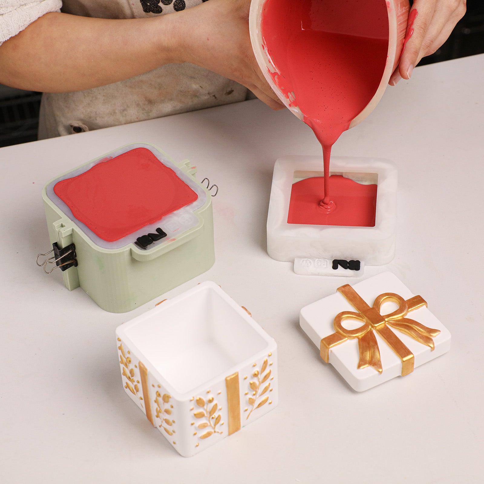 Craft pouring red casting material into Holiday Gift Box Candle Jar two‑part buckle silicone mold, showing finished cast pieces beside the mold - Boowan Nicole