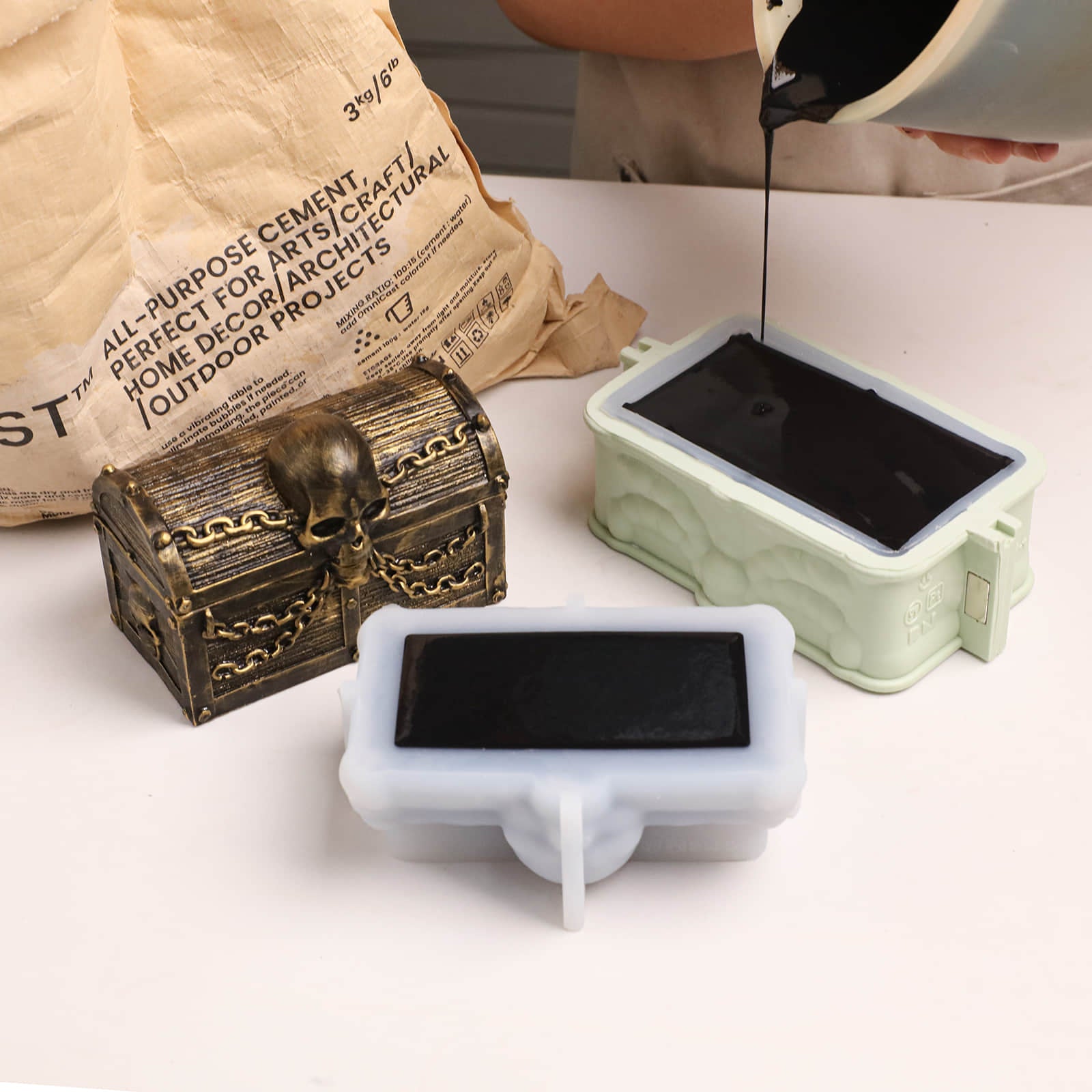 Pouring black casting material into silicone molds for skull treasure chest candle jar, finished sample displayed beside molds - Boowan Nicole