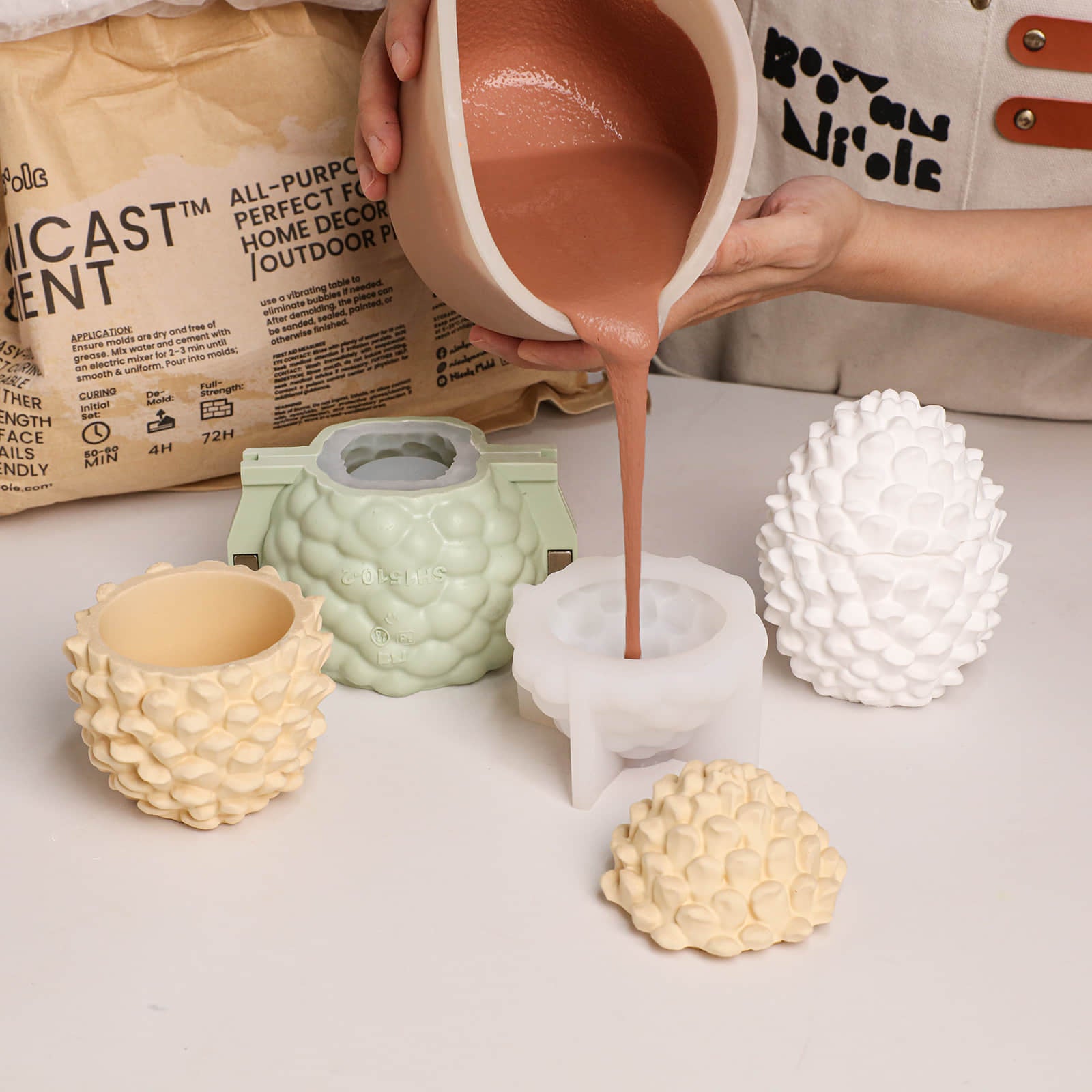 DIY craft scene: pouring pigmented casting compound into Little Woodland Pinecone silicone candle jar molds beside finished candle jar blanks - Boowan Nicole