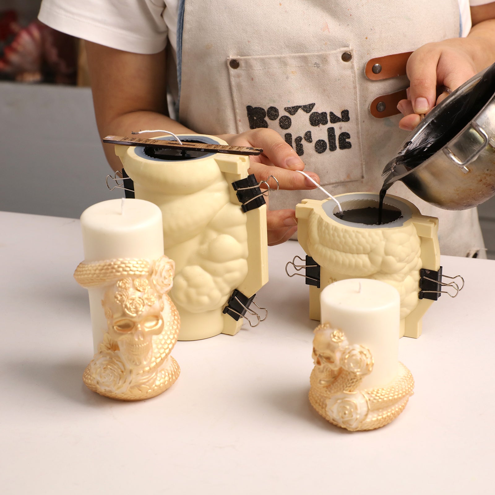Craft workshop scene: pouring black melted wax into Venom & Roses silicone candle molds alongside finished skull snake candle blanks - Boowan Nicole