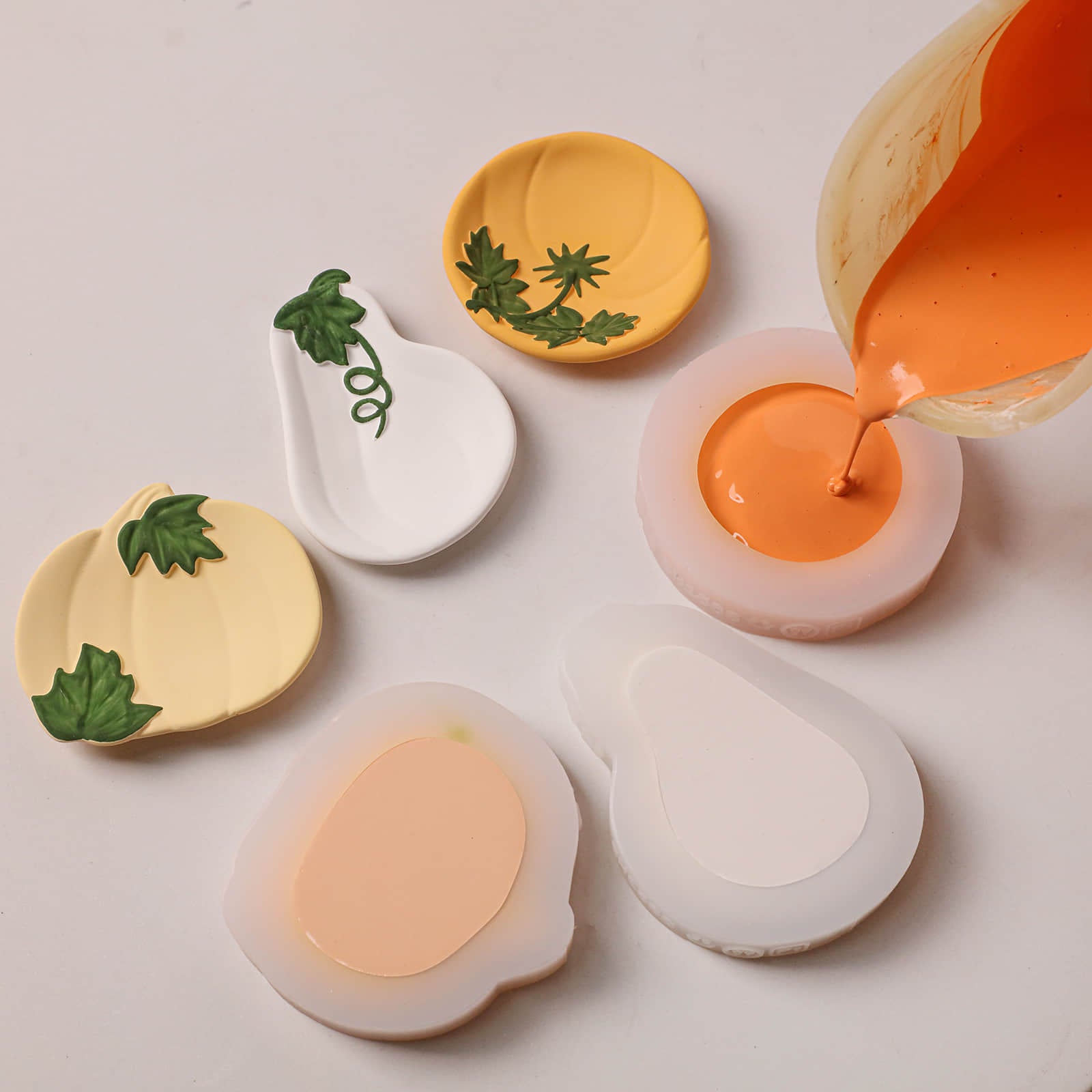Several small decorative dish-like molds or trays in white and pale yellow are arranged on a light surface, with orange liquid being poured into one mold and more orange material visible in another. Some pieces have green leaf or vine motifs.
