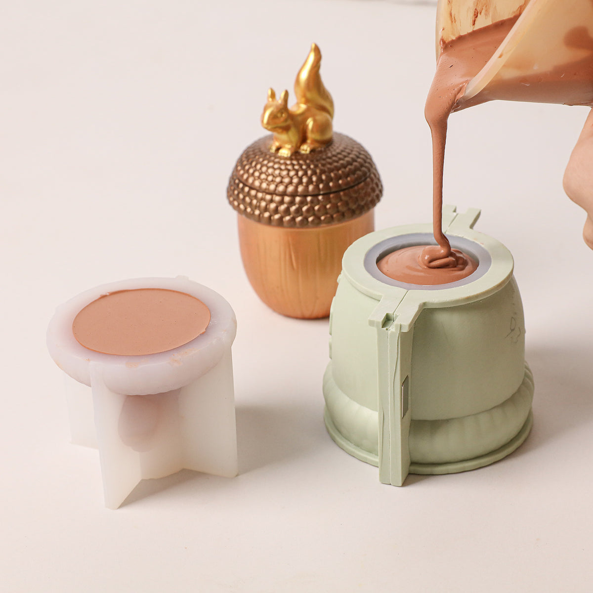 Pouring brown casting material into acorn candle jar silicone mold, finished squirrel acorn candle sample displayed beside the mold - Boowan Nicole