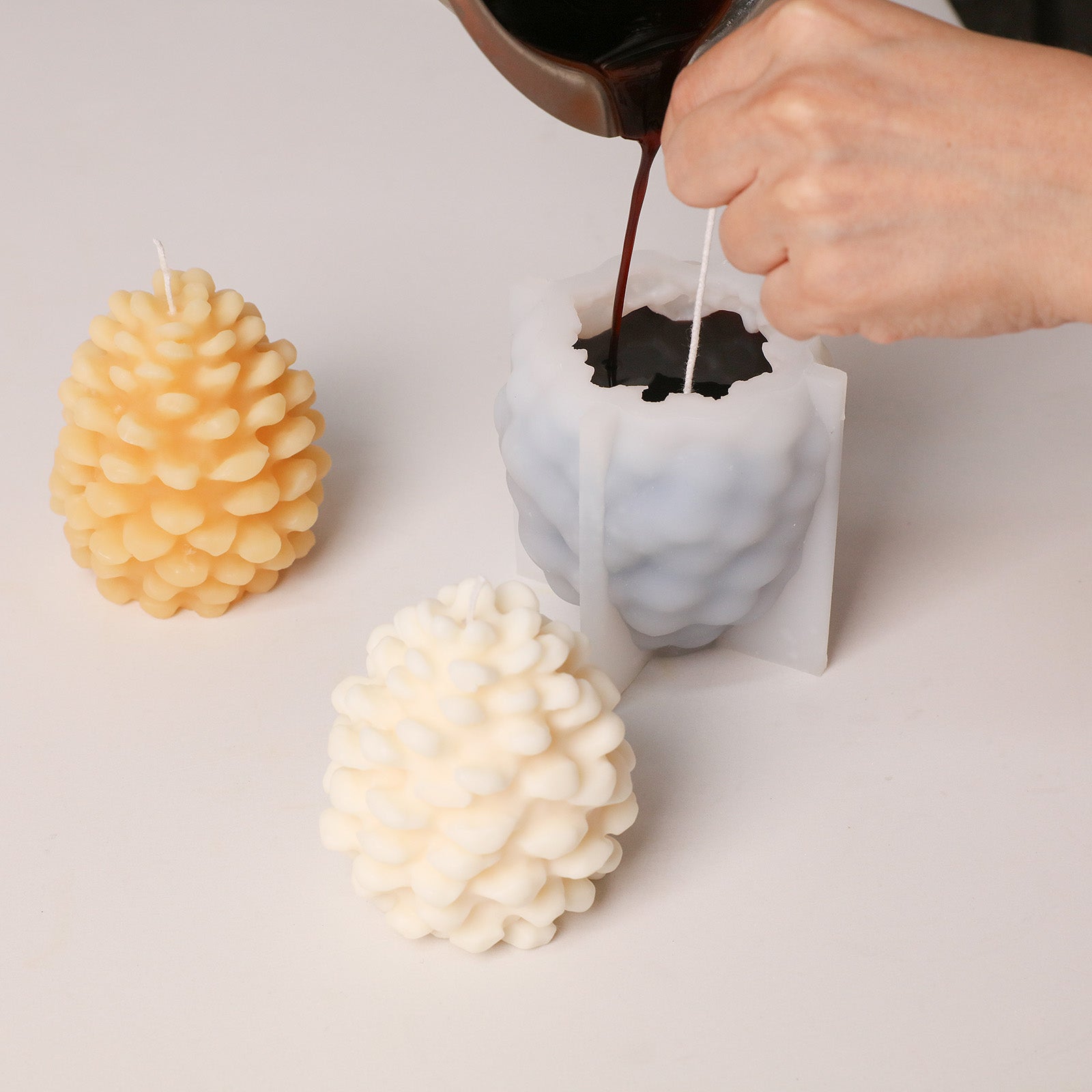 Craft process photo: pouring pigmented wax into winter pinecone silicone candle mold beside finished candle blanks - Boowan Nicole