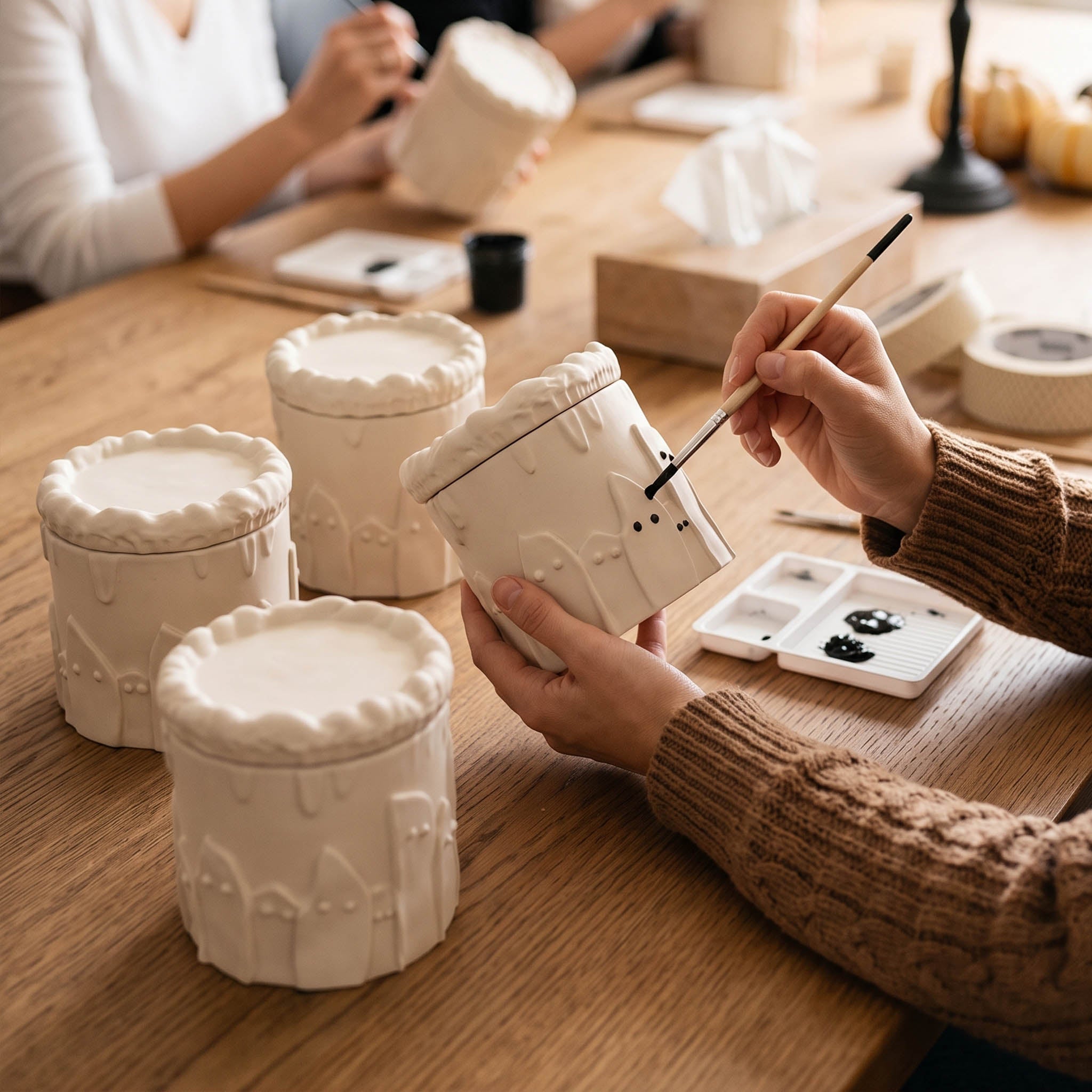 Crafter hand painting small black eyes onto unfinished white ghost cake candle jars during workshop - Boowan Nicole