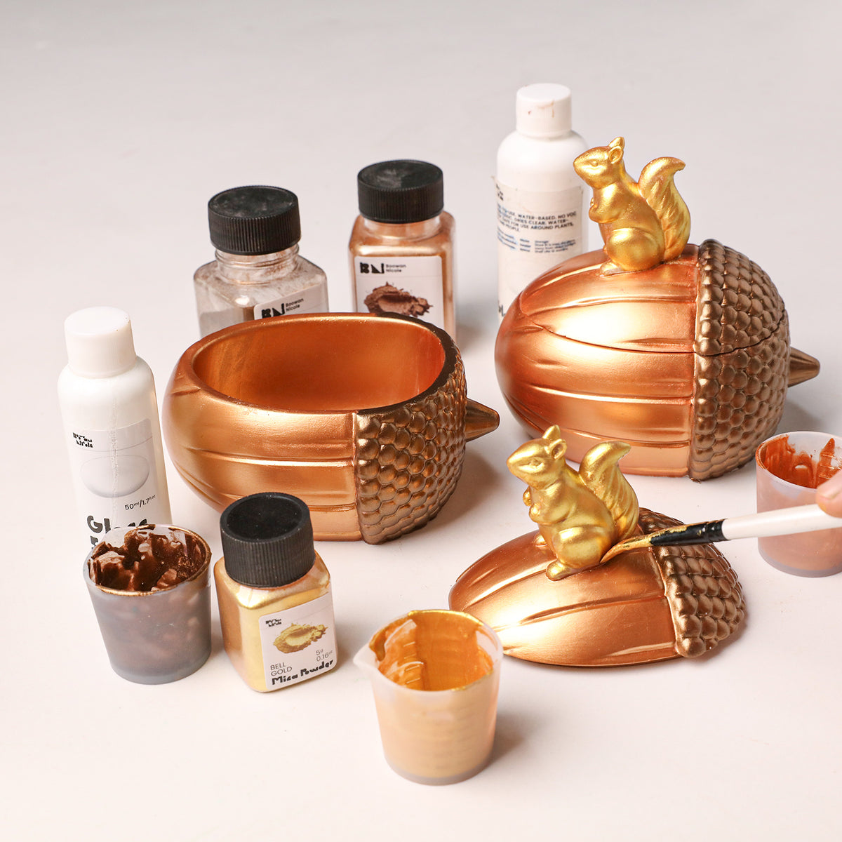 DIY painting golden squirrel nut candle jar with mica powder, assorted pigments and paintbrushes laid out on work table - Boowan Nicole