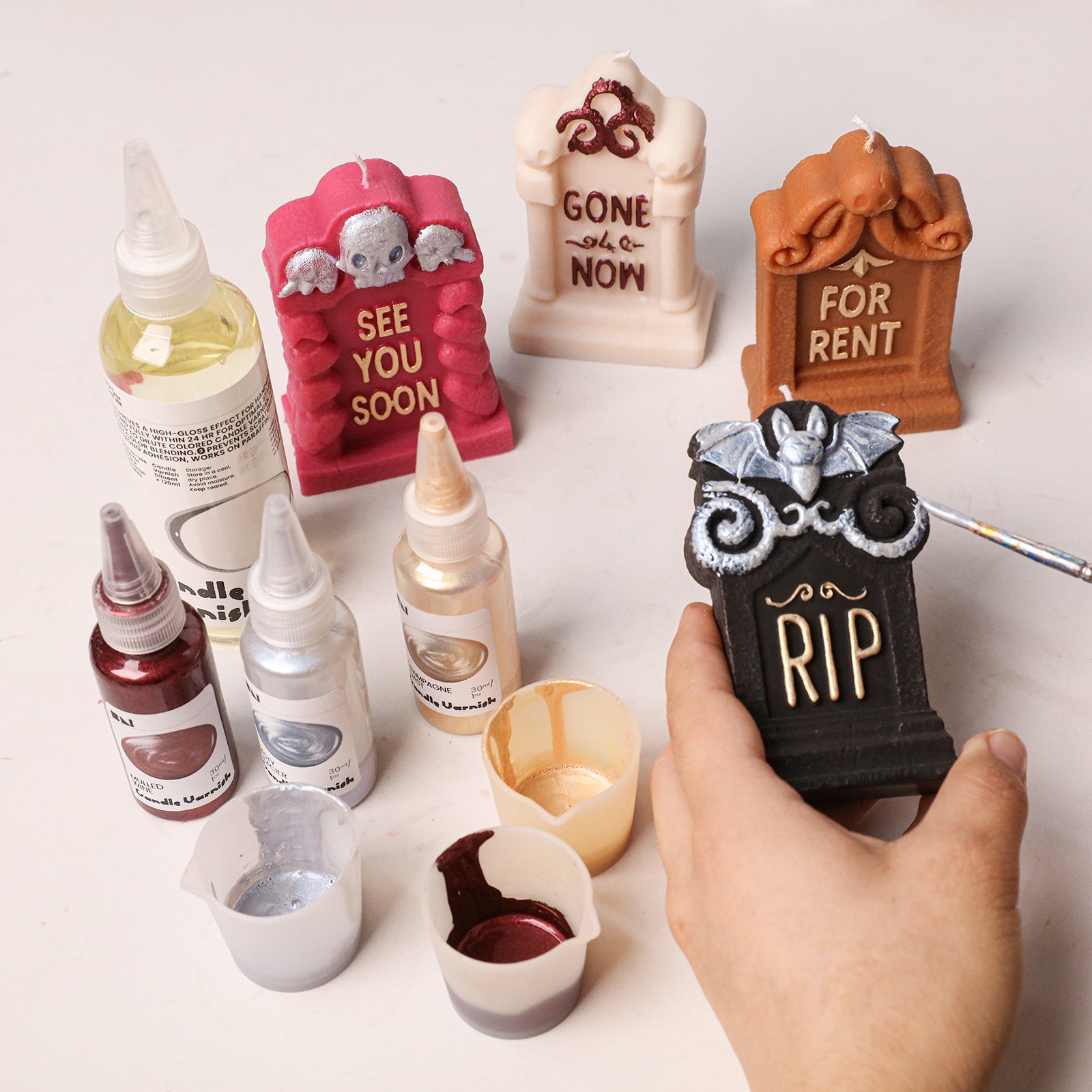 Hand painting metallic detail on black RIP tombstone candle, assorted colored tombstone blanks and candle varnish craft table scene - Boowan Nicole