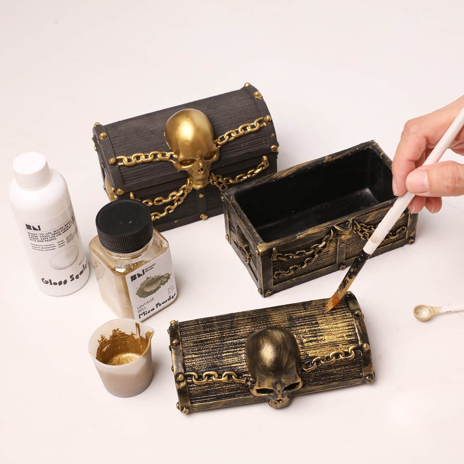 DIY painting unfinished skull treasure chest candle jar blank with mica powder, gloss sealer and painting tools on clean white table - Boowan Nicole