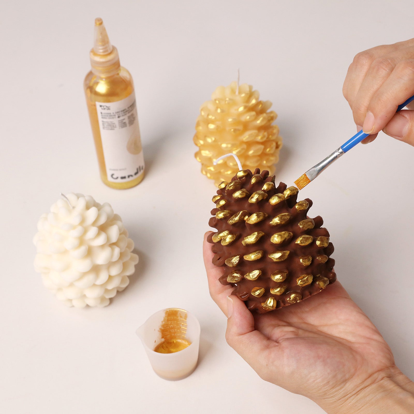 Artisan hand painting metallic gold highlight details on dark brown winter pinecone candle craft table scene - Boowan Nicole