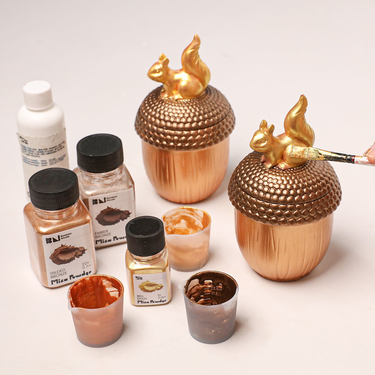 DIY painting acorn squirrel candle jars with gold mica powder and paintbrushes, craft pigment supplies arranged on table - Boowan Nicole