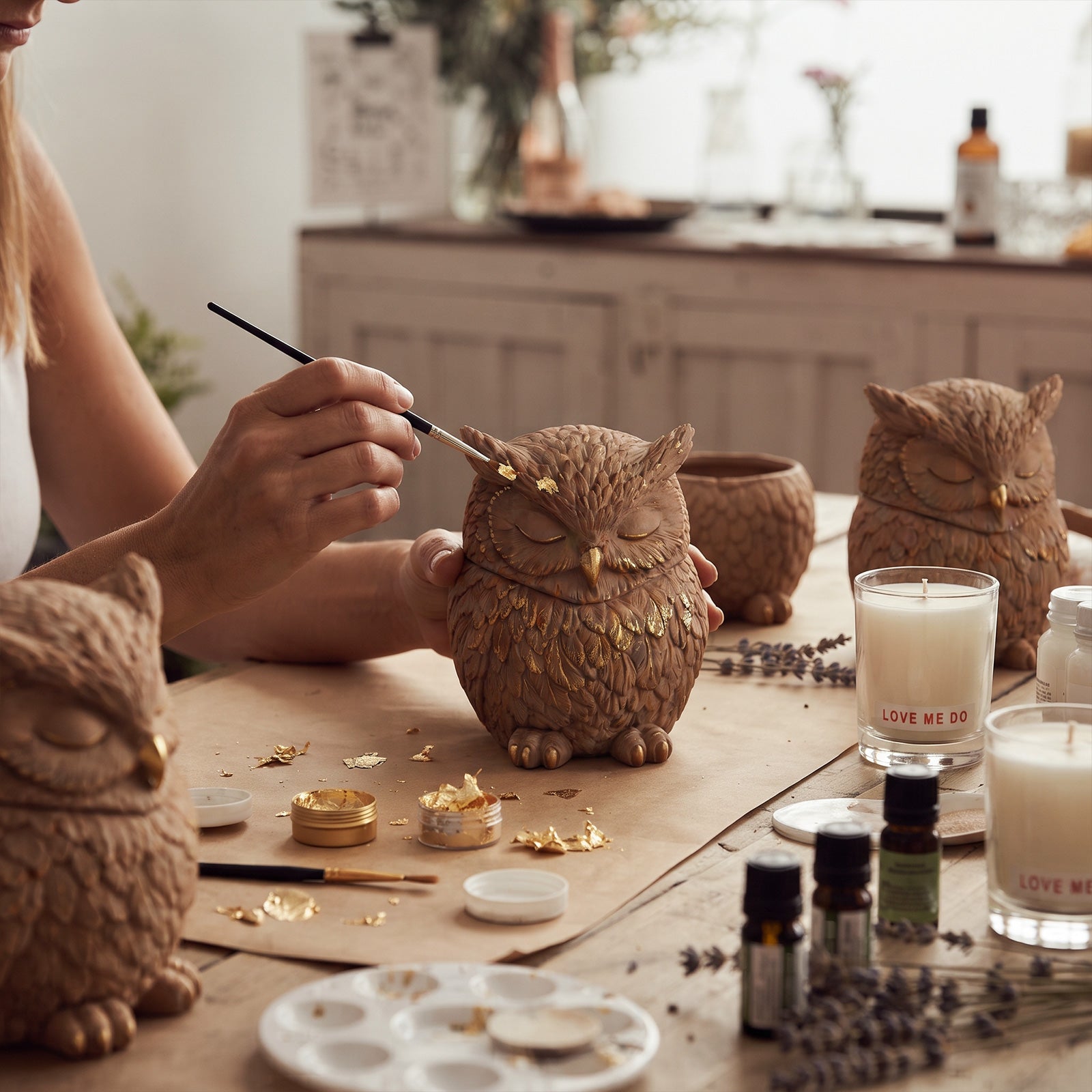 Crafter applying gold leaf gilding decoration onto handmade owl candle jar at craft worktable - Boowan Nicole