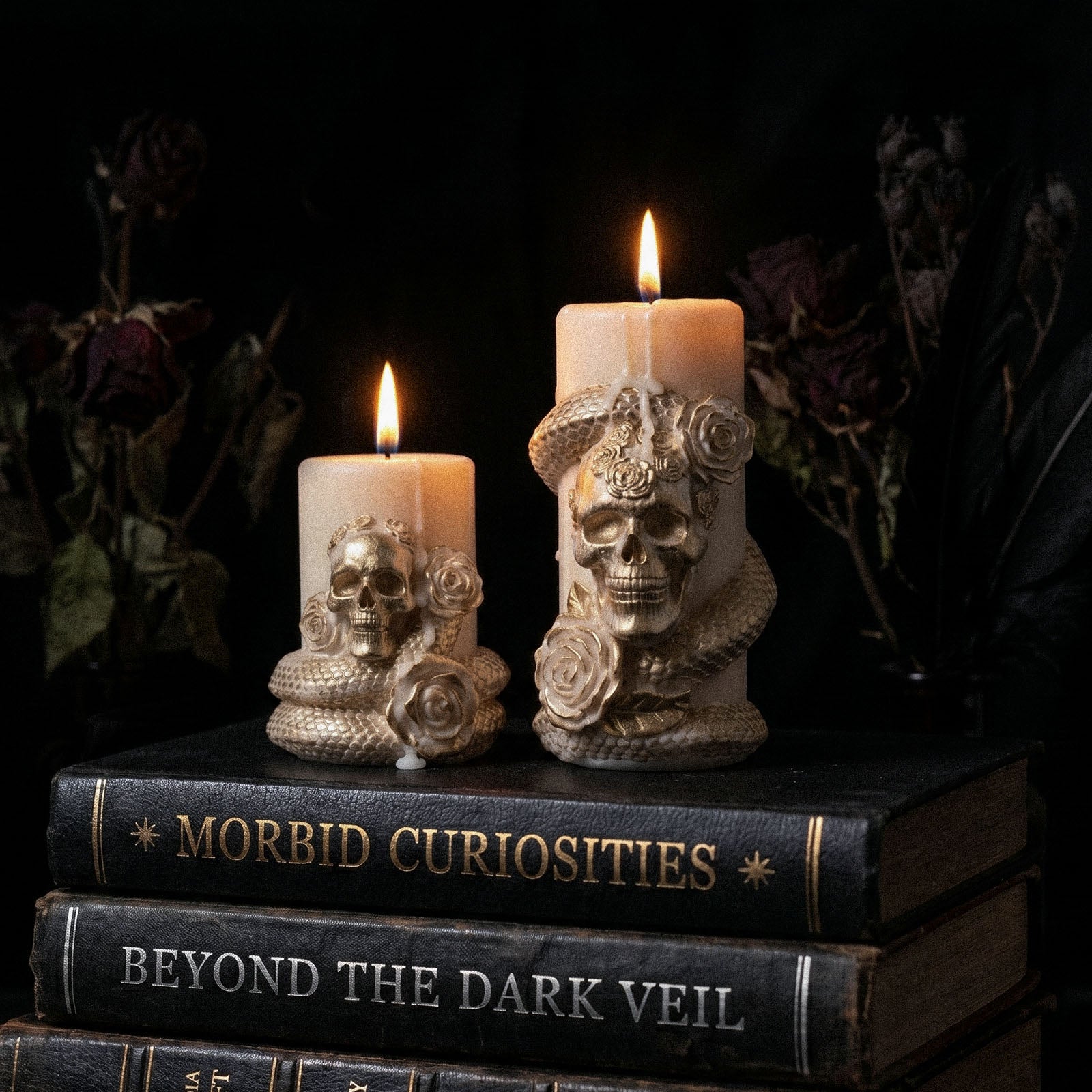 Two sizes lit cream Venom & Roses pillar candles with gold skull serpent rose detailing stacked on vintage goth hardcover books - Boowan Nicole