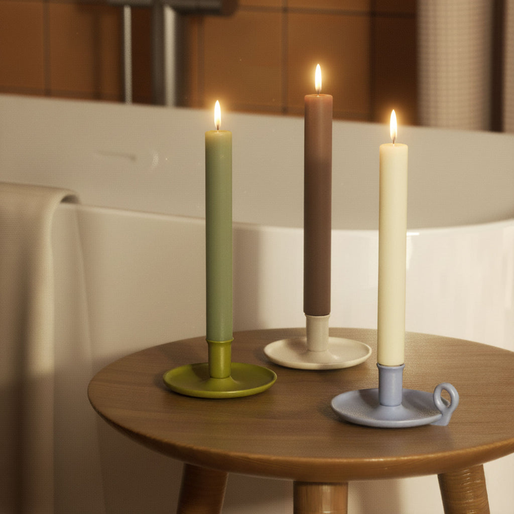 Three lit 9.3" classic taper candles—sage, taupe, and cream—in ceramic holders on a wooden stool by the bath - Boowan Nicole