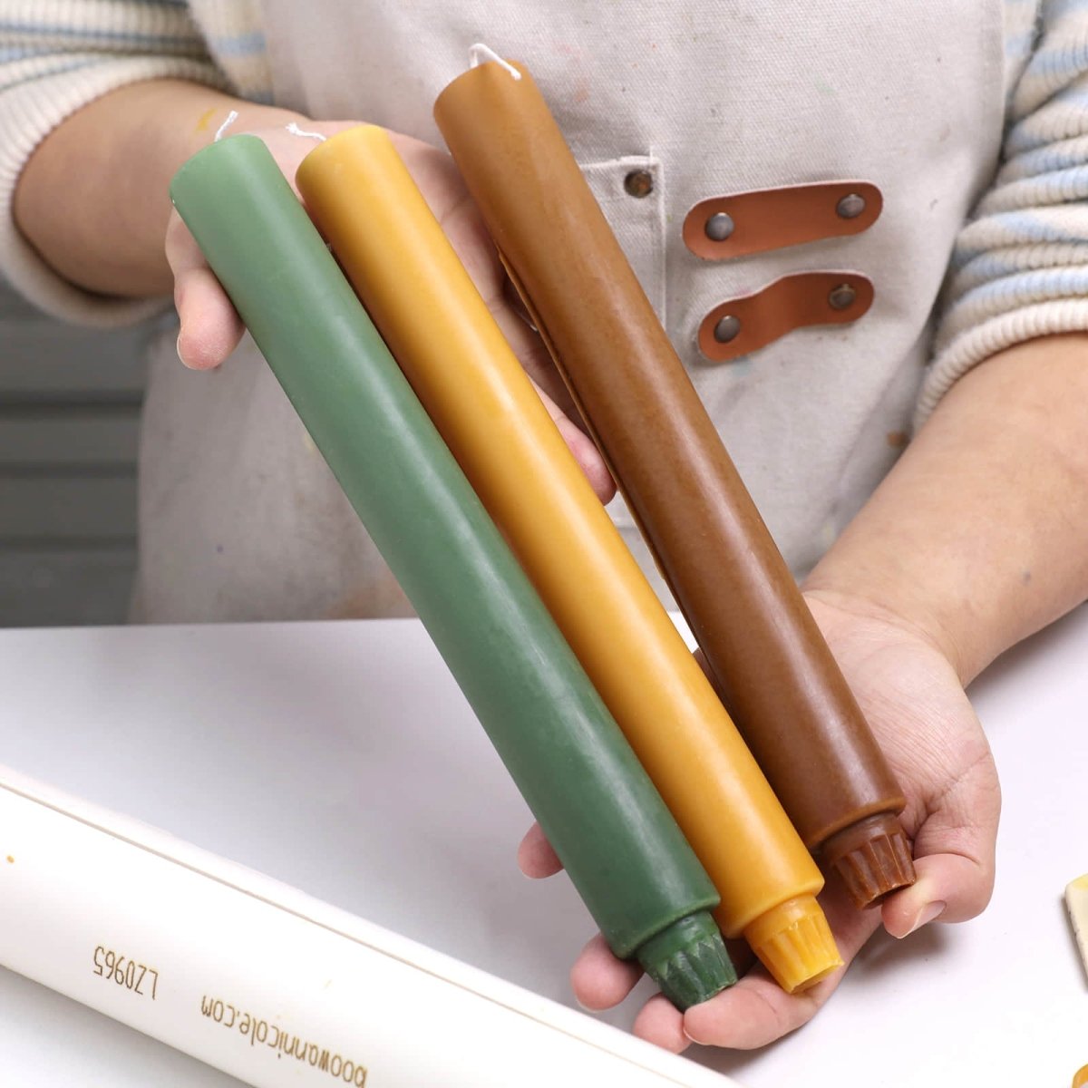Hands holding three 11" classic taper candles—forest green, mustard, and chestnut - Boowan Nicole