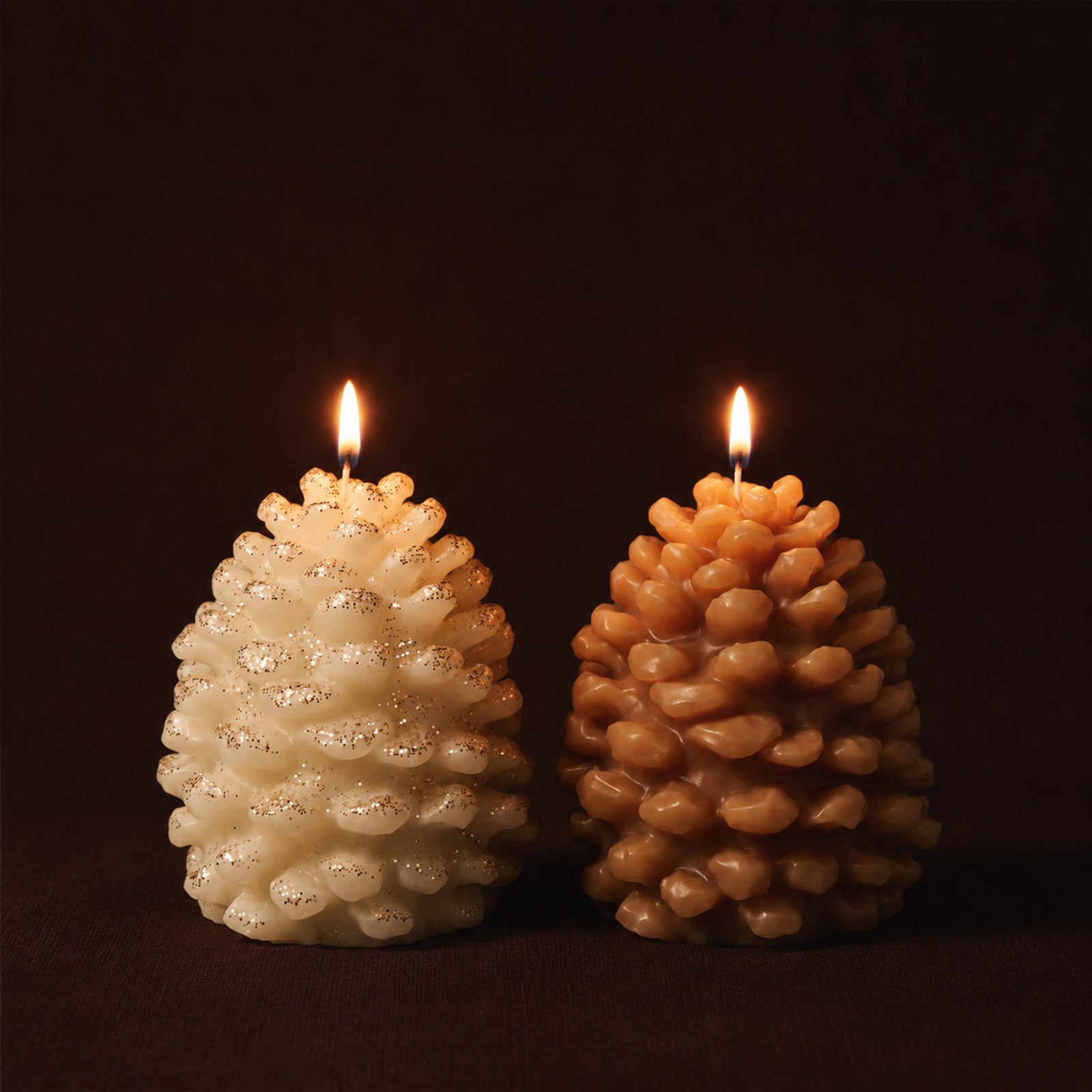 Two lit winter pinecone pillar candles, cream glitter finish and warm caramel tone, dark fabric background moody seasonal product shot - Boowan Nicole
