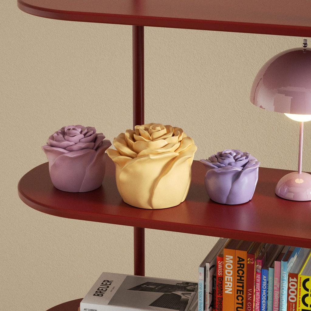 Three rose-shaped pastel candles in different sizes displayed on a red shelf with books and a lamp - Boowan Nicole