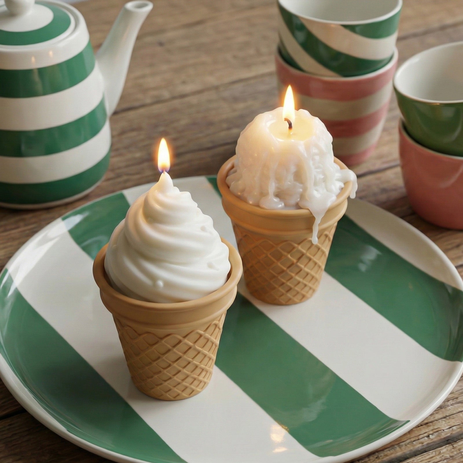 Two lit ice cream cone candles on a green and white striped plate, one with soft-serve style wax and one with a melting scoop design, styled with a matching striped teapot and colorful cups. - Boowan Nicole