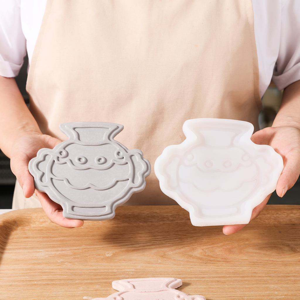 Hand-held display of taupe wavy amphora-shaped coasters and white silicone molds, designed by Boowan Nicole.