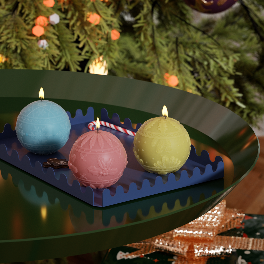 Three festive Christmas ball candles in blue, pink, and yellow, with intricate embossed designs, glow warmly on a decorative tray under the holiday tree.