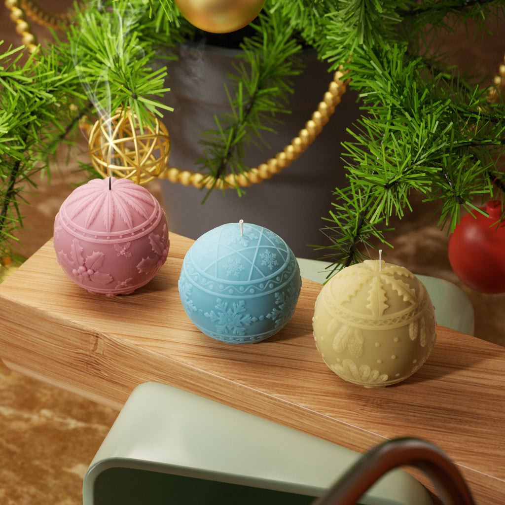 Three festive ball candles with 3D snowflake designs in red, blue, and yellow, displayed under a Christmas tree on a wooden surface.