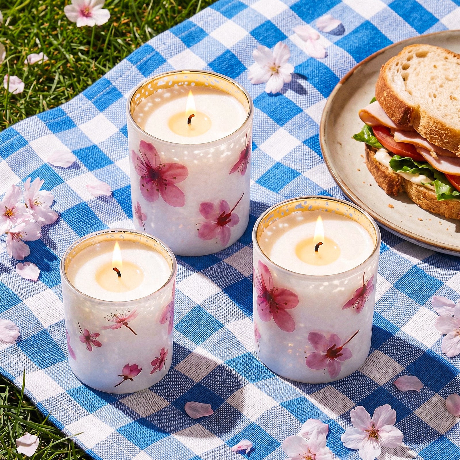 Three Burning Sakura Printed Candle Cups Laid on Blue Gingham Picnic Blanket With Fresh Petals and Sandwich - Boowan Nicole