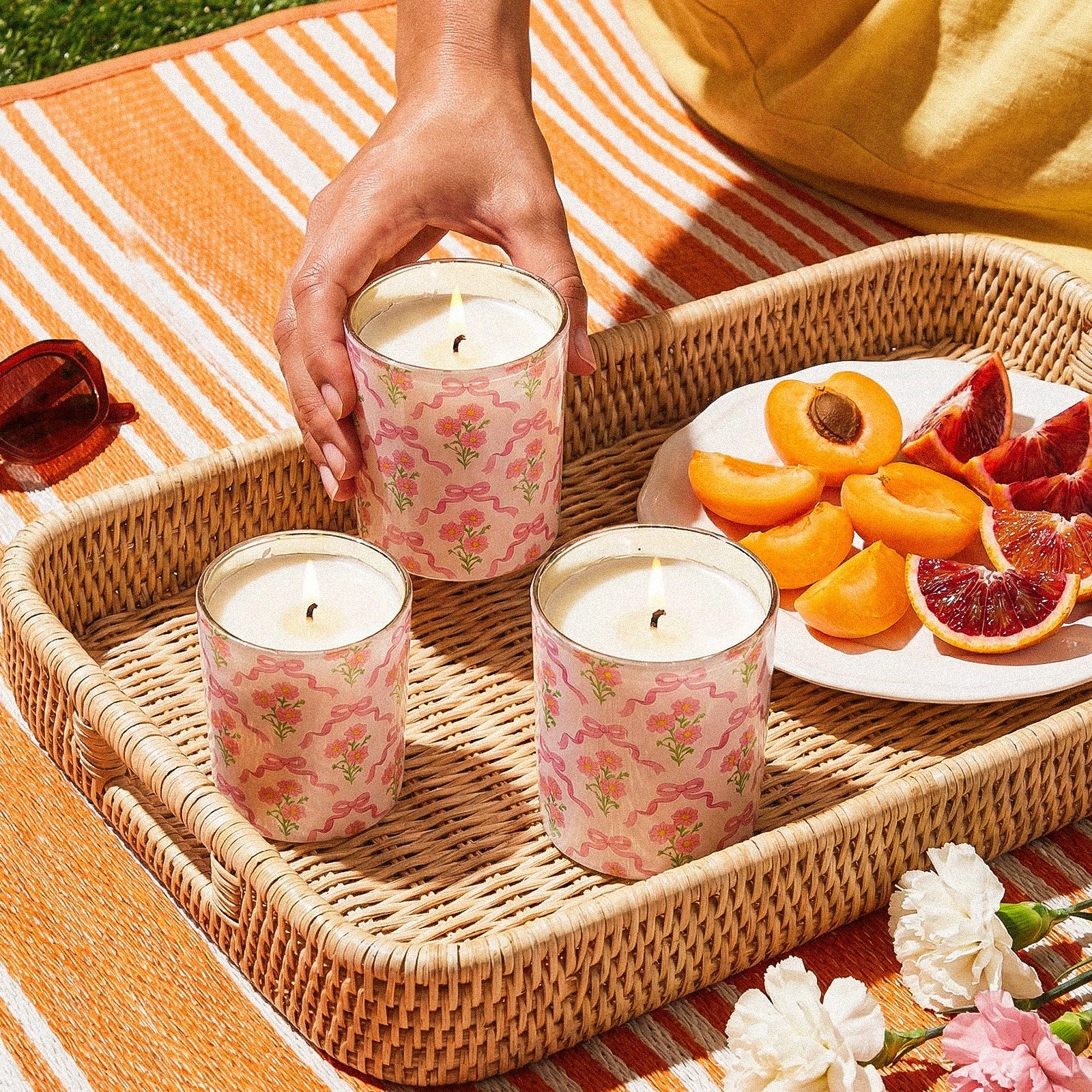 Three Burning Pastel Floral Bow Printed Candle Cups Rested on Rattan Picnic Tray With Fresh Stone Fruits and Outdoor Striped Fabric - Boowan Nicole