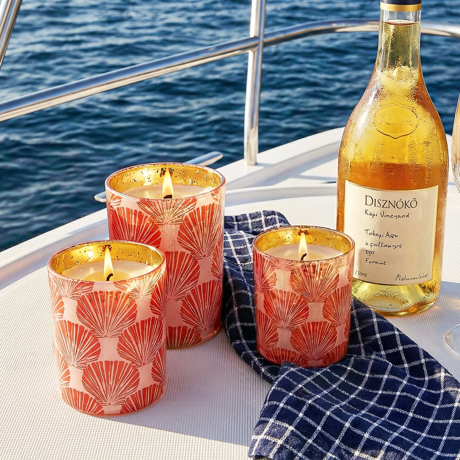 Three Burning Seashell Print Candle Cups Laid on Dark Blue Plaid Fabric Beside White Wine on Yacht Deck - Boowan Nicole