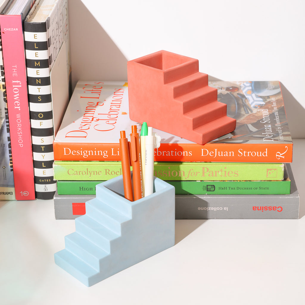 There are three ballpoint pens inserted in the blue Stairway Pen Holder, and an orange-red Stairway Pen Holder is placed on the book next to it -Boowan Nicole