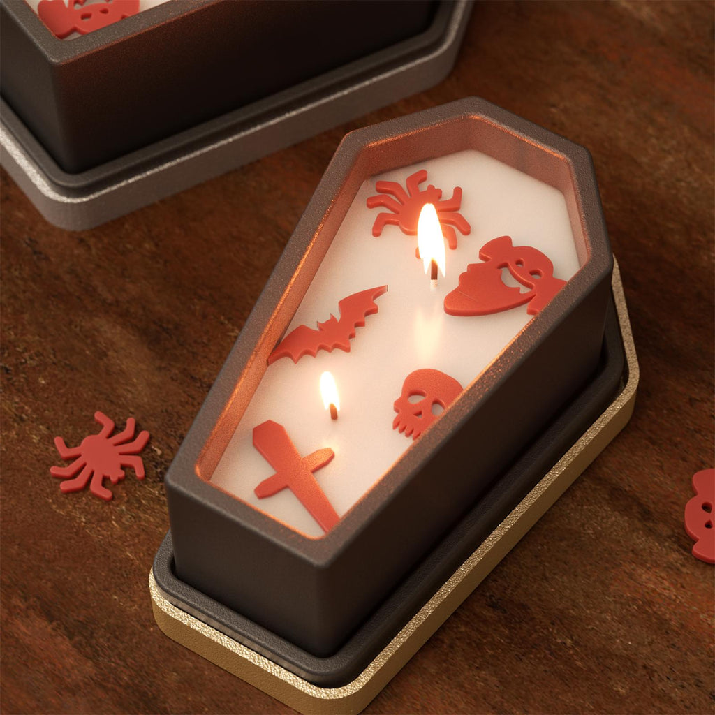 Coffin-shaped Halloween candle with embedded red wax decorations including spider, skull, bat, and ghost face, lit with three flames on a rustic tabletop - Boowan Nicole