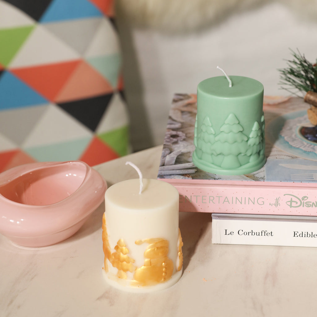 The green pillar-shaped Christmas-themed embossed candle on the table and the Christmas snowman embossed candle painted with golden mica powder on the book next to it - Boowan Nicole