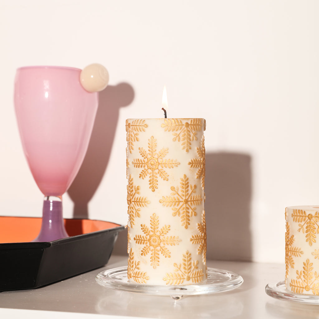 The long white cylindrical snowflake relief candle that is being lit is placed in a tray on the table, blending harmoniously with the background.