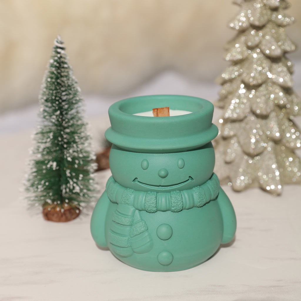 A green Cheery Snowman's Winter Glow Candle Jar sits on a tabletop with Christmas decorations behind it - Boowan Nicole