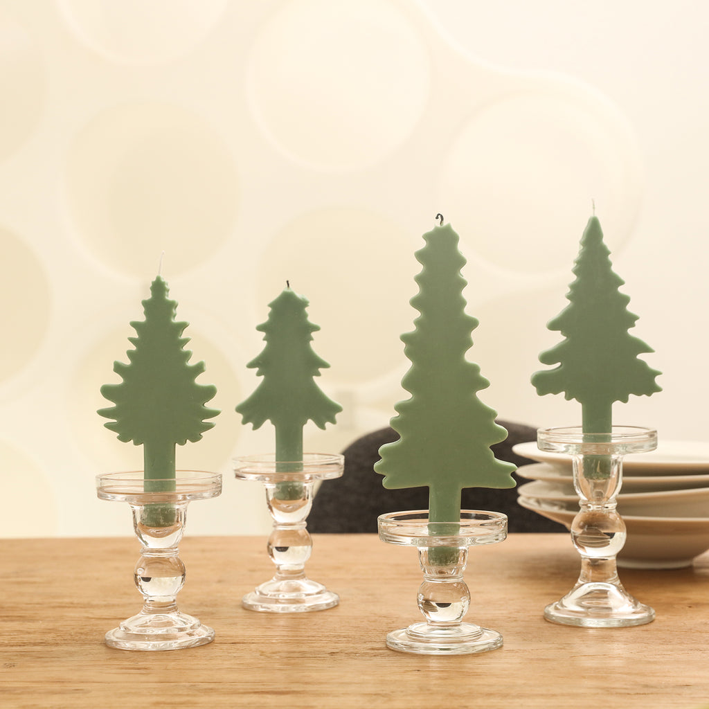 Four Christmas tree-shaped candles of different shapes and sizes are placed on the crystal candle holders on the table.