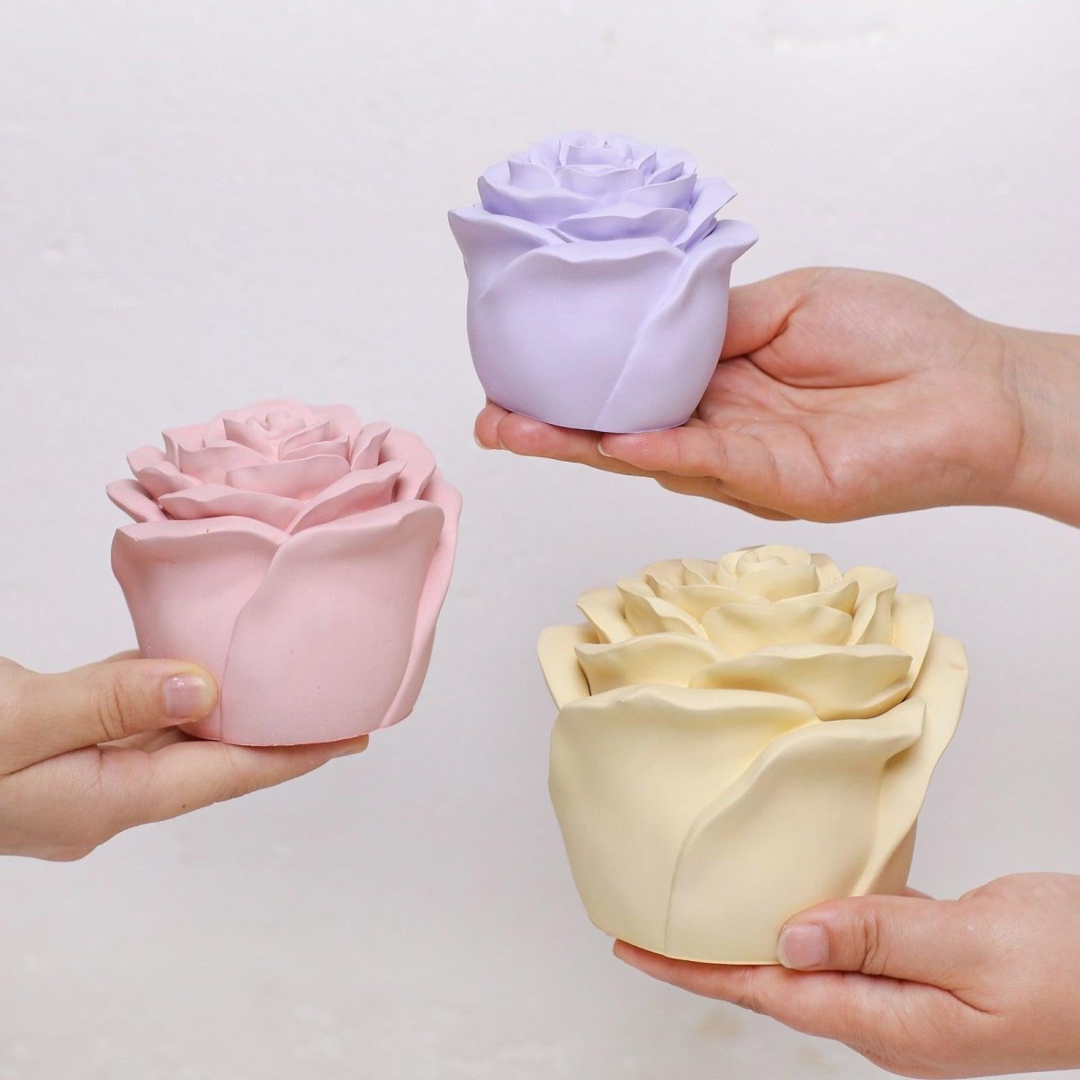 Three pastel-colored rose-shaped candle jars in pink, purple, and yellow held in hands - Boowan Nicole