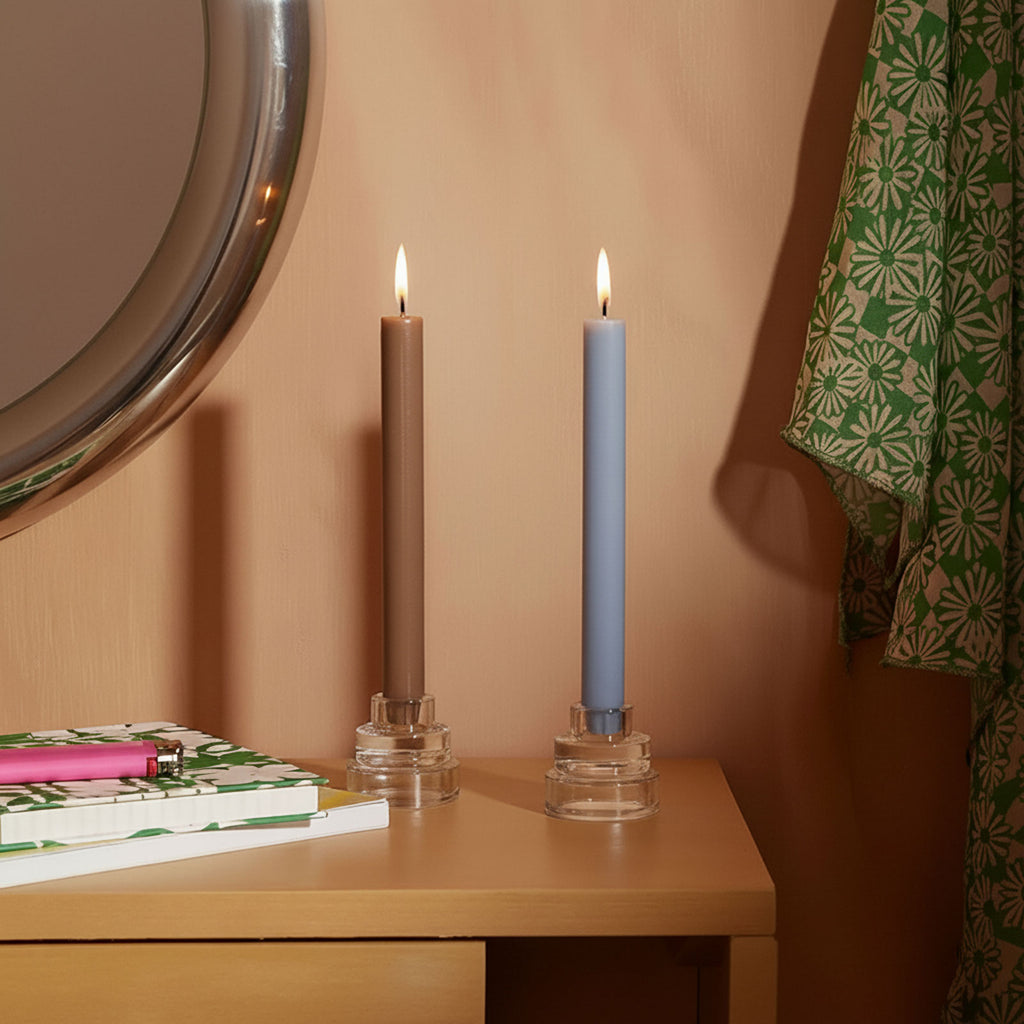 Two lit 9.3" classic taper candles—taupe and light blue—in clear glass holders on a bedside table - Boowan Nicole