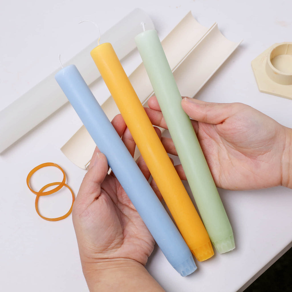 Three pastel 9.3" classic taper candles—blue, yellow, and mint green—with fluted bases, shown in hand after demolding from a silicone mold - Boowan Nicole