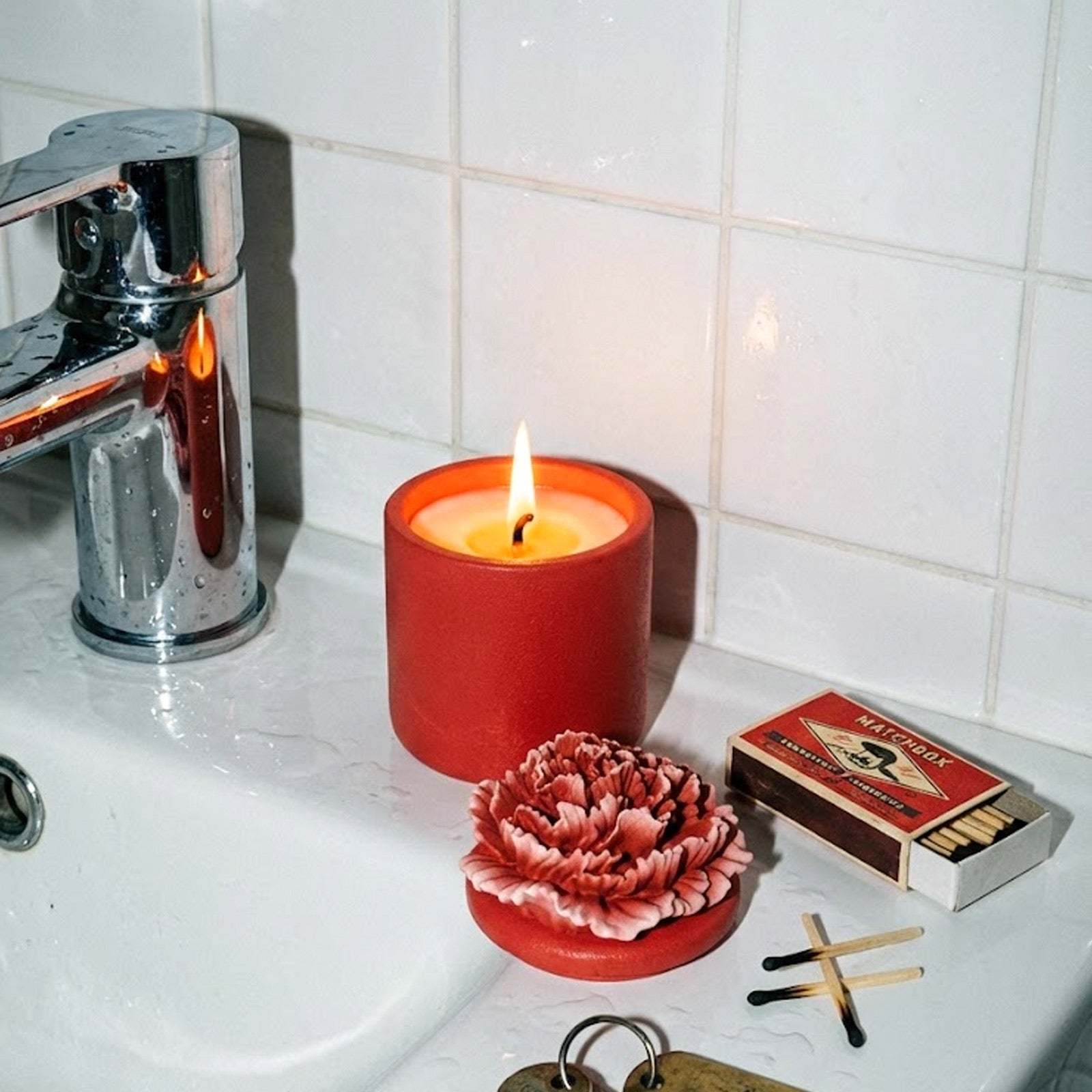 Lit red carnation candle jar with a separate carnation lid on a bathroom sink -Boowan Nicole