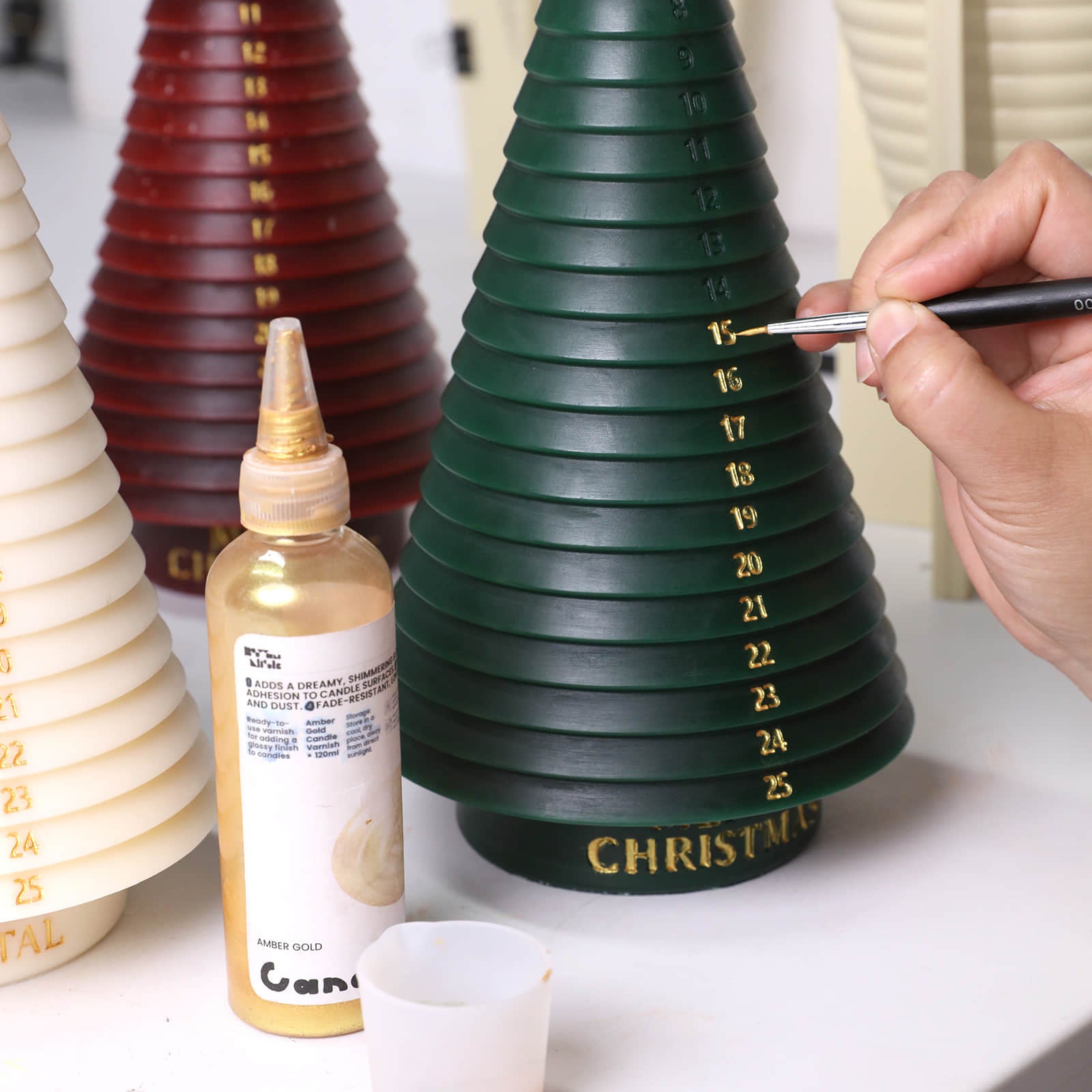 Painting gold countdown numbers on a green Advent tree candle; gold pigment bottle nearby - Boowan Nicole