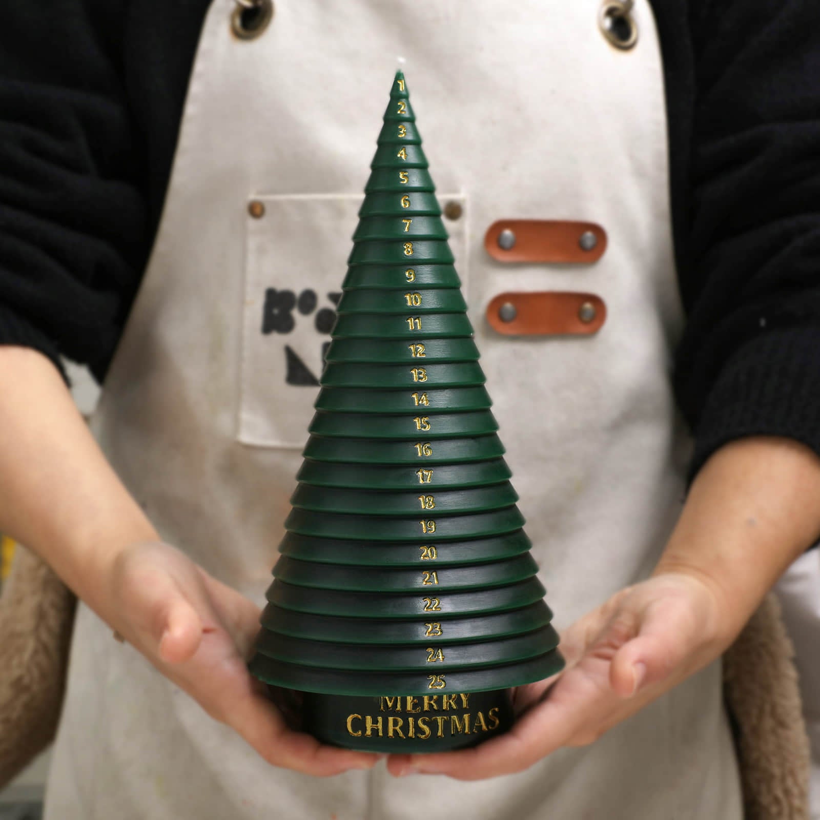 Hands holding a green Advent countdown Christmas tree candle with gold numbers and “Merry Christmas” base - Boowan Nicole