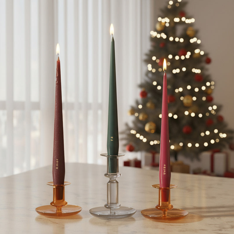 Three taper candles—burgundy, green, and berry—in amber and clear glass holders on a table, Christmas tree bokeh behind; gold text “Amor,” “Fé,” and “União.” - Boowan Nicole