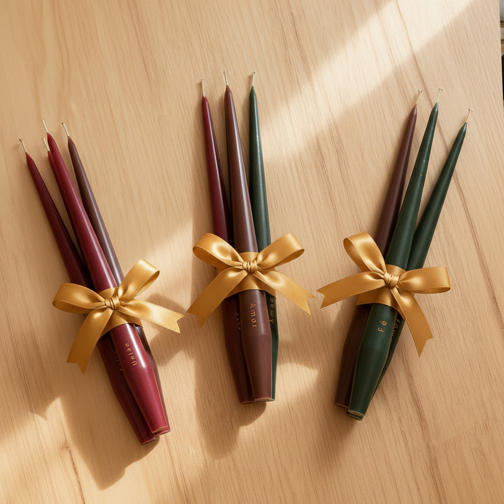 Gift-ready taper candle bundles tied with gold bows—burgundy, brown, and forest green—arranged on a wooden surface - Boowan Nicole